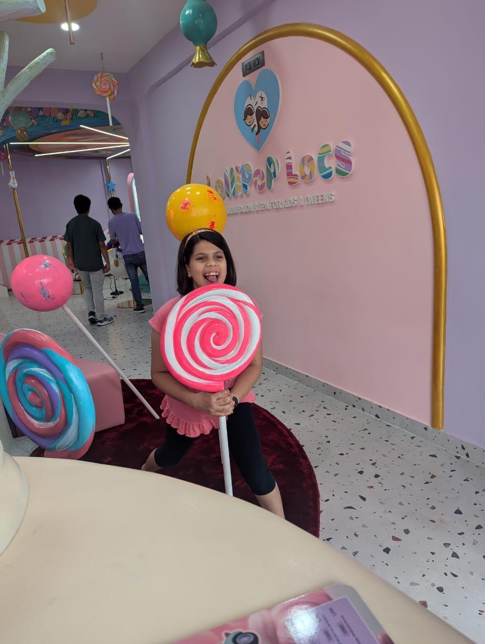 Young girl posing with a giant pink and white lollipop prop in front of the Lollipop Locs salon logo